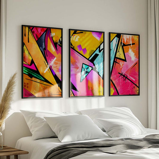 Three abstract paintings in yellow, pink, black, and blue colors with text elements hanging on the wall above a white bed with gray throw blanket and wooden nightstand with potted plant in a modern bedroom. Artwork by Kali Nuevo.