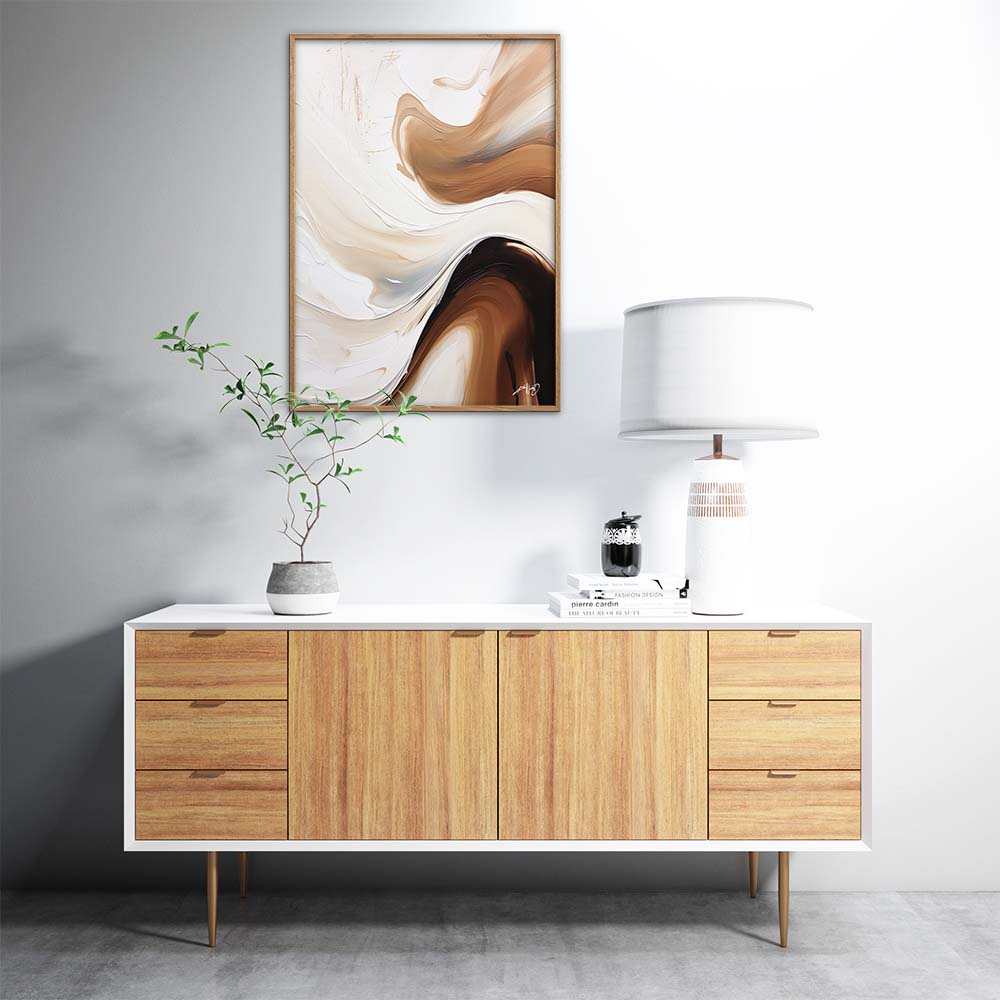 A brown, white, and black abstract painting with fluid, organic shapes on a white wall above a wooden stand with a lamp and a plant.