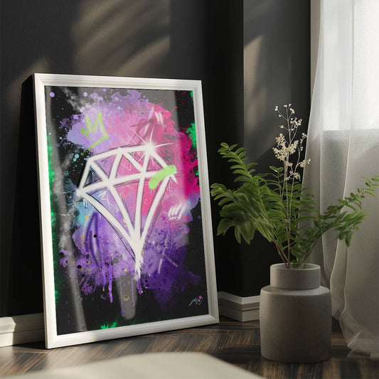 A framed artwork of a white diamond with colorful splashes in the background leaning against a dark wall next to a potted plant and a window with a sheer curtain in a bright room. Artwork by Kali Nuevo.
