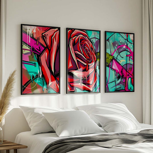 Three framed pieces of abstract artwork in red, green, pink, and blue colors with a rose motif hanging on the wall above a white bed with dark throw blanket and wooden nightstand with potted plant in a modern bedroom. Artwork by Kali Nuevo.